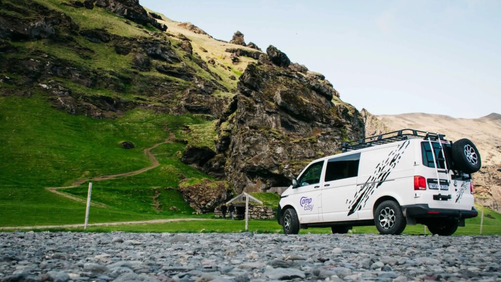 A white camper van parked on rocky ground near a lush green hillside, with a winding path and rugged cliffs in the background.
