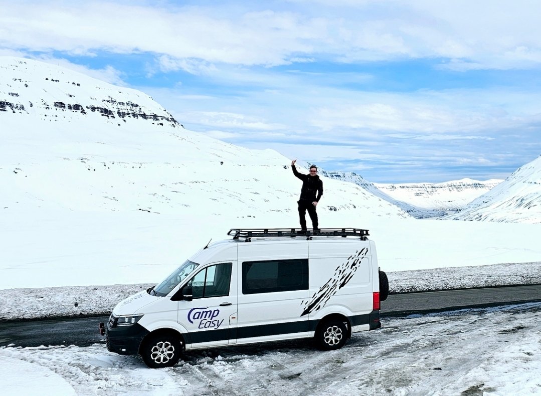 A person stands on the roof rack of a white camper van, parked on a snowy road surrounded by vast snowy mountains. The person is dressed in black, waving happily under a clear blue sky.