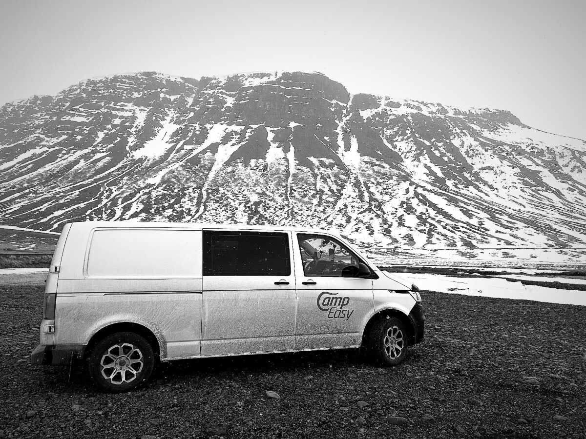 A white camper van parked on a gravel surface in front of a large, snow-patched mountain. The surroundings appear cold and desolate, reflecting a stark yet majestic winter landscape