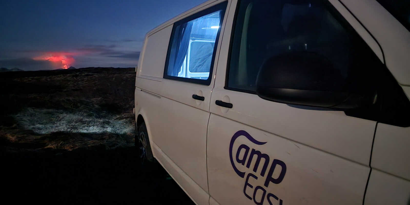 Nighttime view of a CampEasy campervan parked in a rugged Icelandic landscape, with a vibrant volcanic eruption glowing in the background, highlighting unique travel experiences in Iceland.