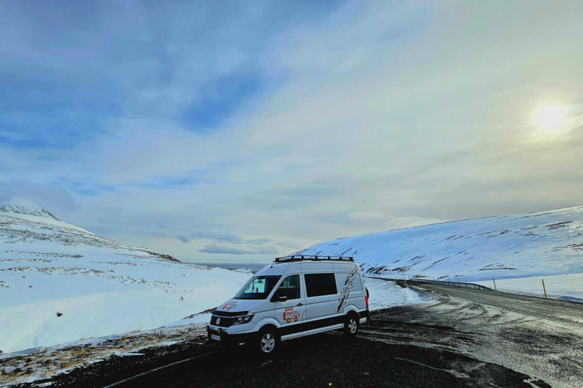 A white campervan parked on a roadside during winter, showcasing expansive snowy mountain landscapes under a clear blue sky in Iceland.