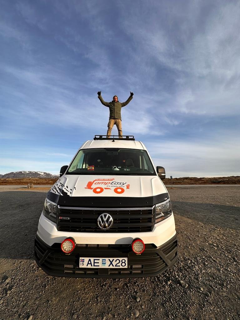 Joyful man standing with arms raised on top of a CampEasy campervan, set against a vast Icelandic landscape under a clear blue sky.
