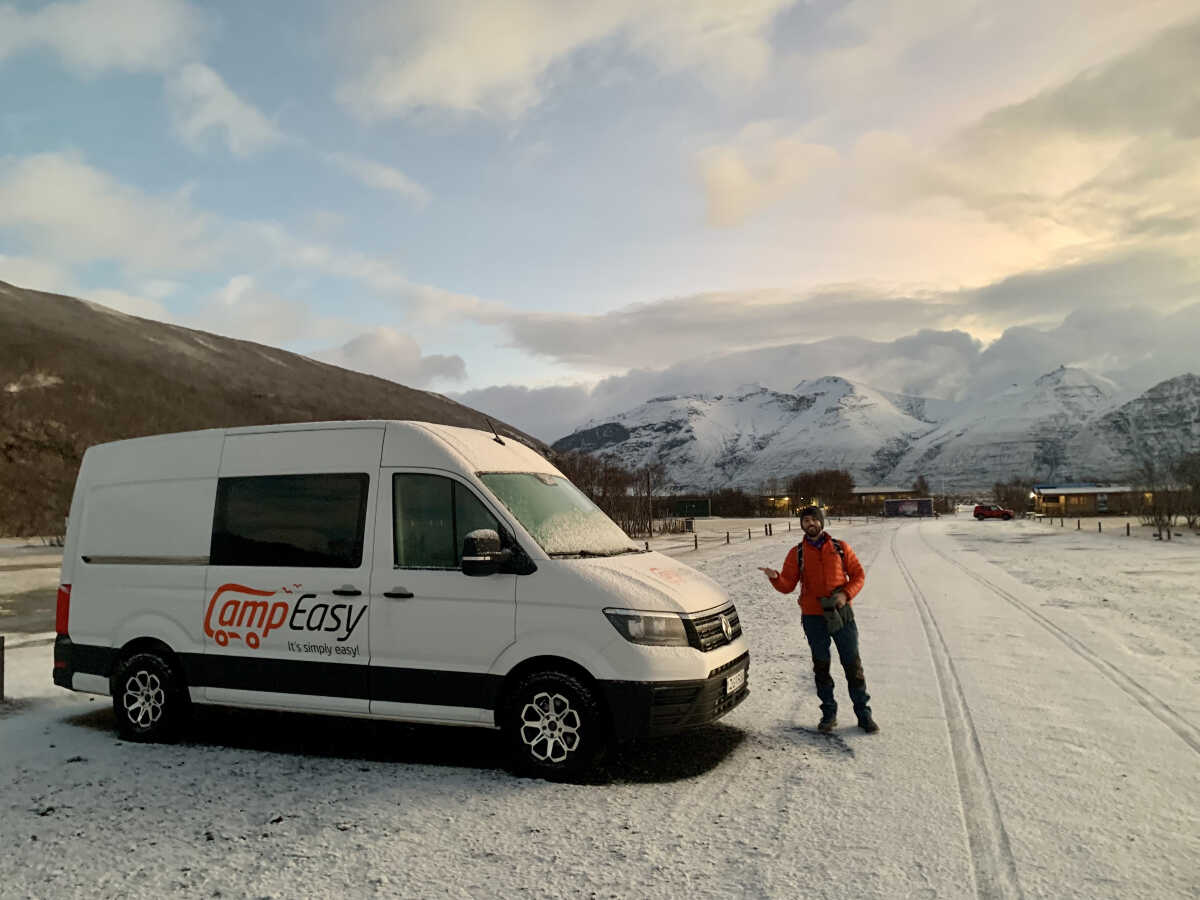 A person in an orange jacket stands next to a CampEasy campervan on a snow-covered road in Iceland, with majestic snow-capped mountains in the background during a bright winter day.