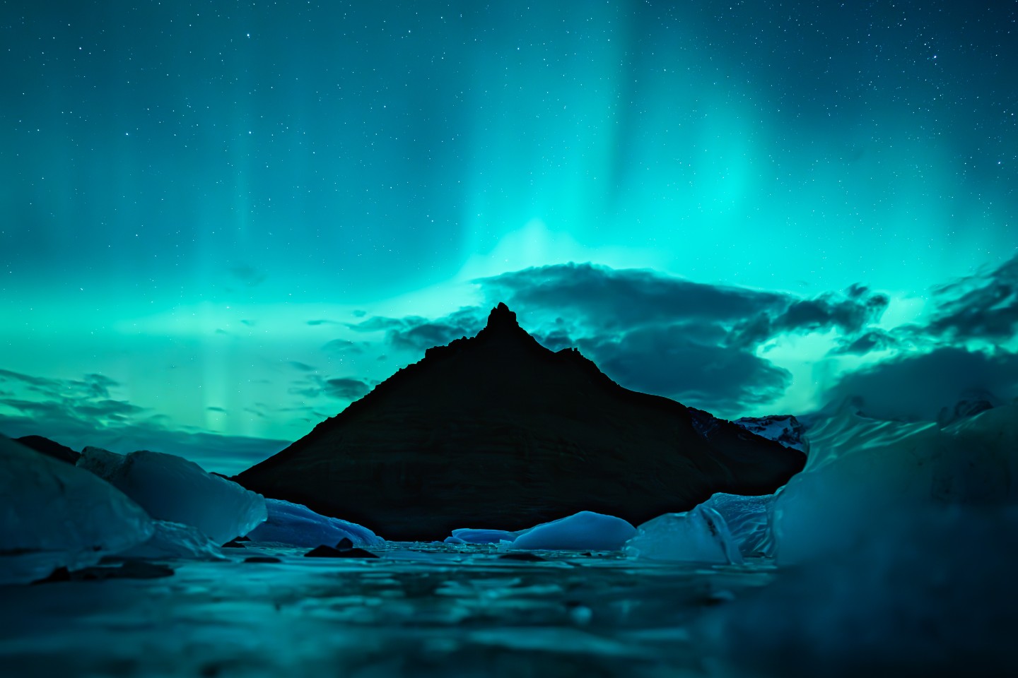 Northern lights glowing above a sharp mountain peak in Iceland, with floating ice in the foreground.