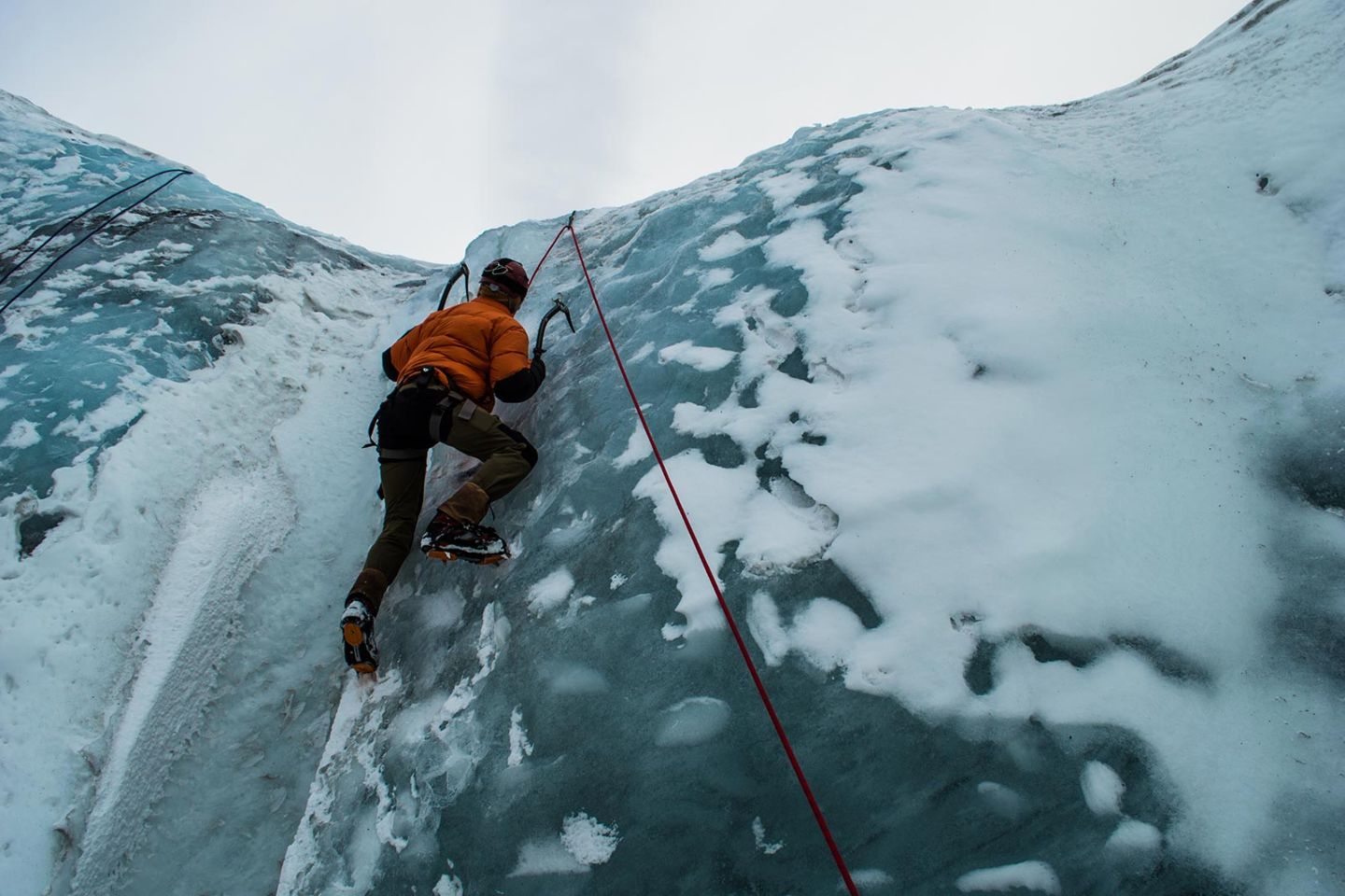 A climber wearing an orange jacket and crampons ascends a steep, icy glacier using ice axes and a safety rope.