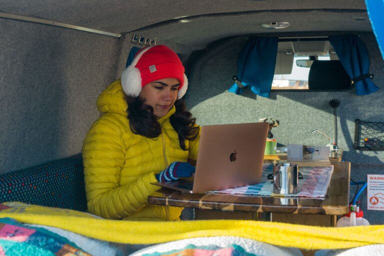 A person in a yellow jacket works on a laptop inside a cozy van, surrounded by colorful blankets and a small table with a mug.