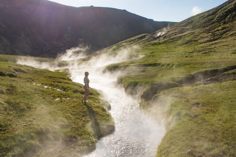 Person standing by a steaming hot spring river in a green Icelandic valley, surrounded by mist and hills.