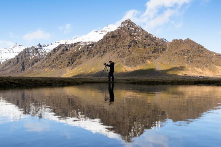 Photographer standing by a reflective pond with a picturesque mountain backdrop under a clear sky.