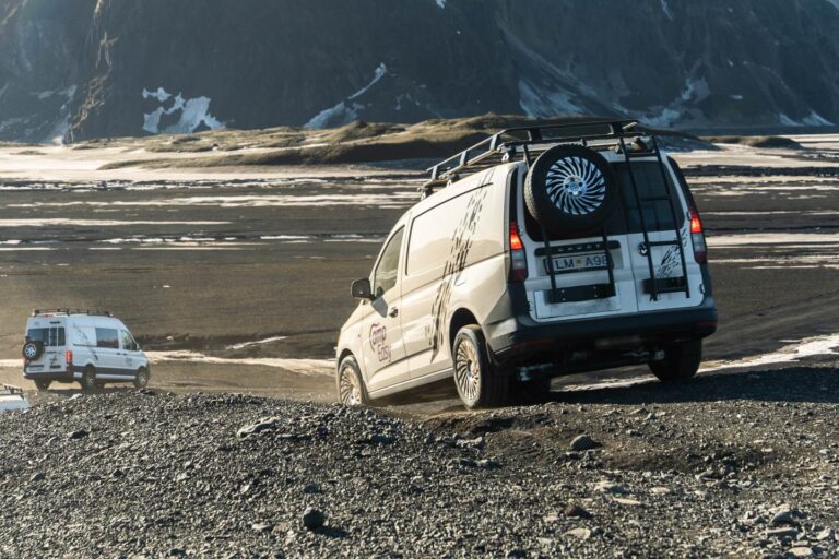 Two white Camp Easy camper vans driving on a rugged dirt road in a remote Icelandic landscape with mountains in the background.