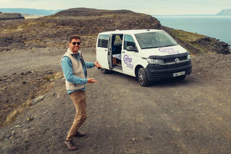 Man standing by white campervan with "CampEasy" logo in Iceland, near cliff and ocean with rugged landscape.