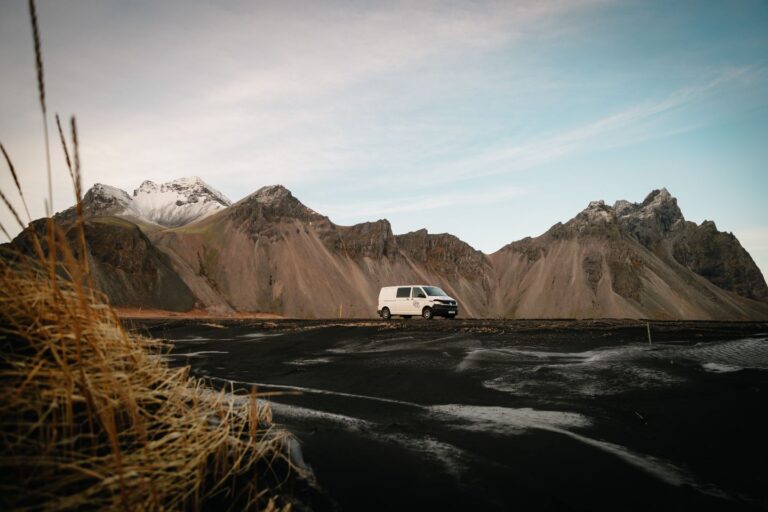 Camper van parked on the gravel road next to the black sand and the huge mountains in the background.