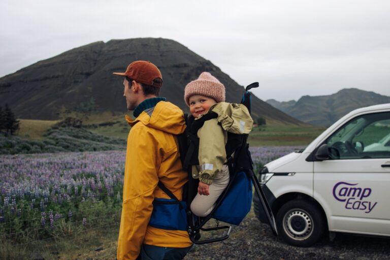Father carrying a smiling child in a hiking backpack near a campervan in Iceland, surrounded by mountains and purple wildflowers.