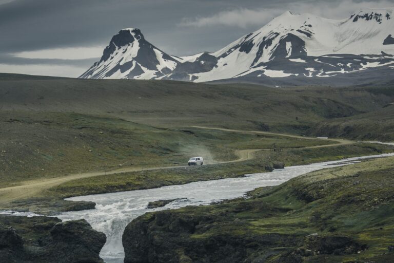 Campervan driving on a remote dirt road in Iceland, surrounded by rugged green landscapes and snow-capped mountains.