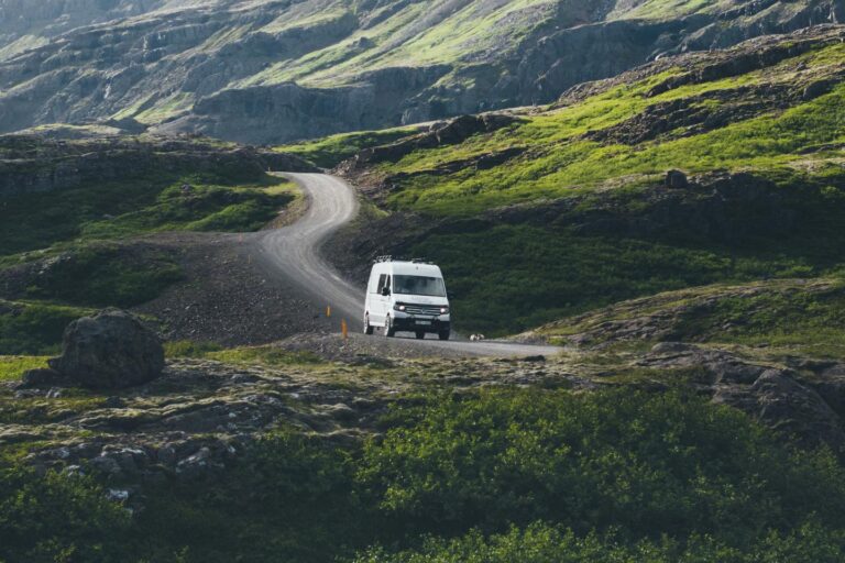 White campervan driving on a winding gravel road through green hills and rocky terrain in Iceland.