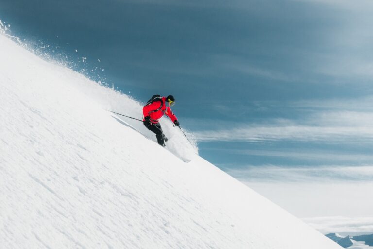 A skier in a red jacket and black pants carves down a steep snowy slope, kicking up powder against a clear blue sky.