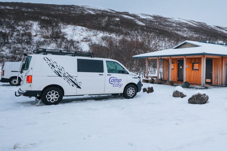 A white Camp Easy camper van parked near a wooden cabin in a snowy Icelandic landscape