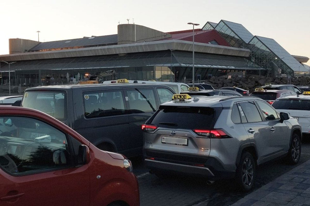 The image shows a row of taxis lined up outside Keflavรญk International Airport in Iceland. The modern airport building with glass and geometric architectural features serves as the backdrop. The scene appears to be during early morning or evening hours, as indicated by the low sunlight.