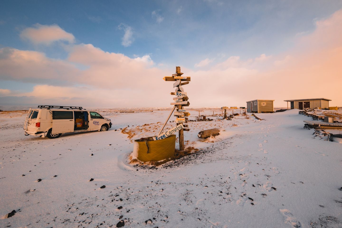 A snowy landscape features a white camper van and a signpost with directional signs, flanked by simple cabins under a colorful sky.