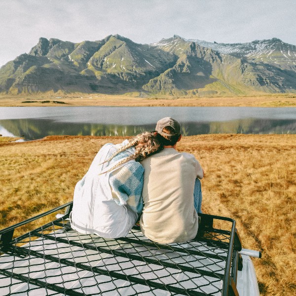Two people are sitting on the roofrack of white campervan and watching the fjord and the mountains