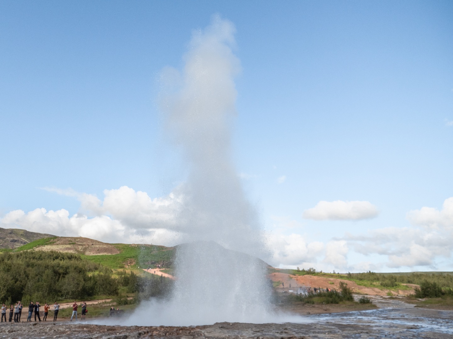 A powerful geyser erupts against a clear blue sky surrounded by green hills. Tourists stand in awe at a distance, capturing the scene on cameras.