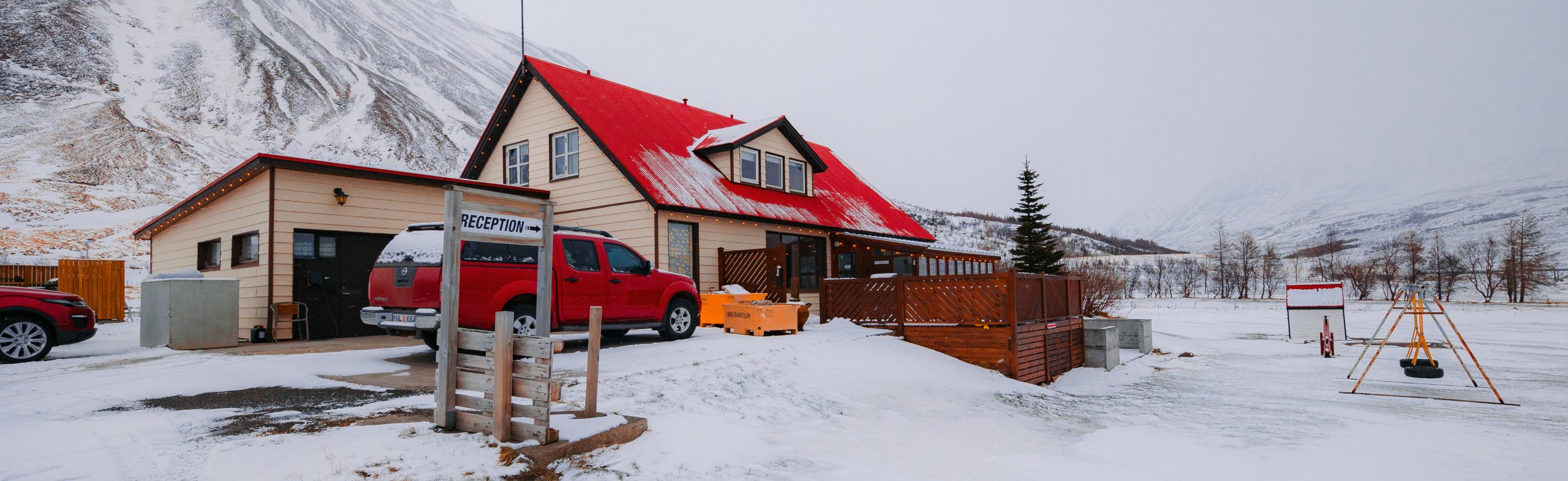 A rustic house with a vibrant red roof stands in a snowy landscape. A red truck is parked nearby, a sign reads "Reception," and snowy mountains loom in the background.