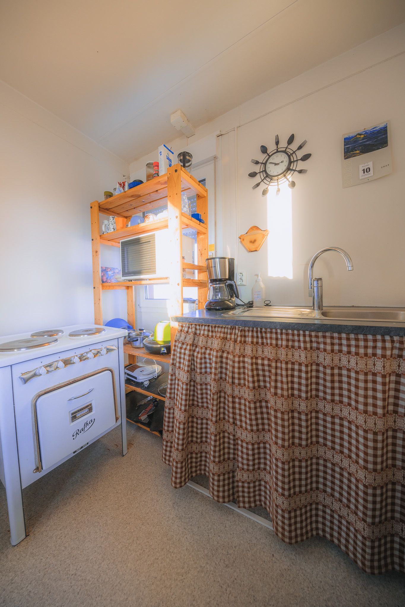 Compact, cozy kitchen with a vintage stove, wooden shelves, checkered skirted sink, wall clock, and light through a window.