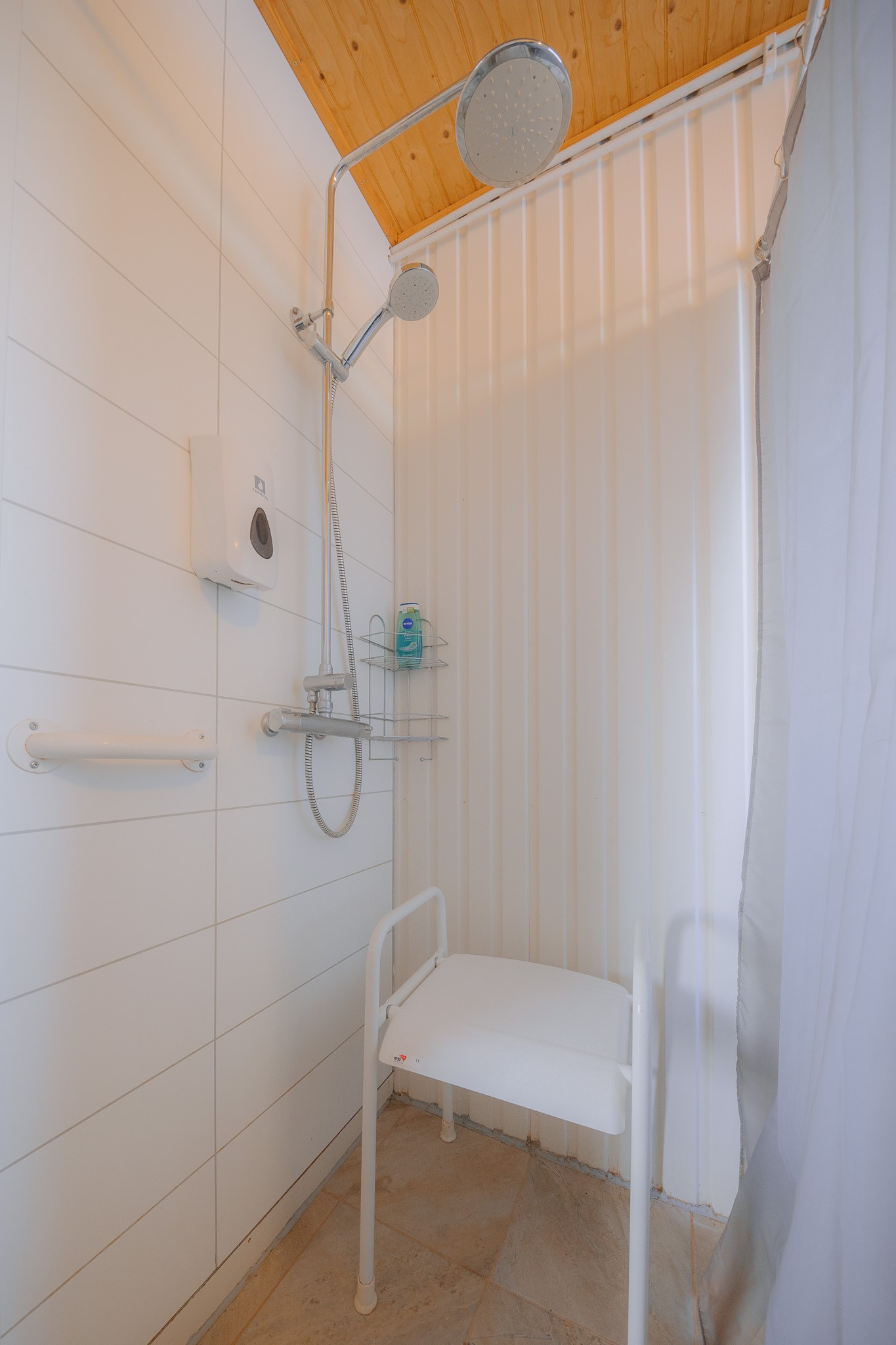 Accessible shower with a wall-mounted seat, grab bars, and a showerhead. Light wooden ceiling and white walls.