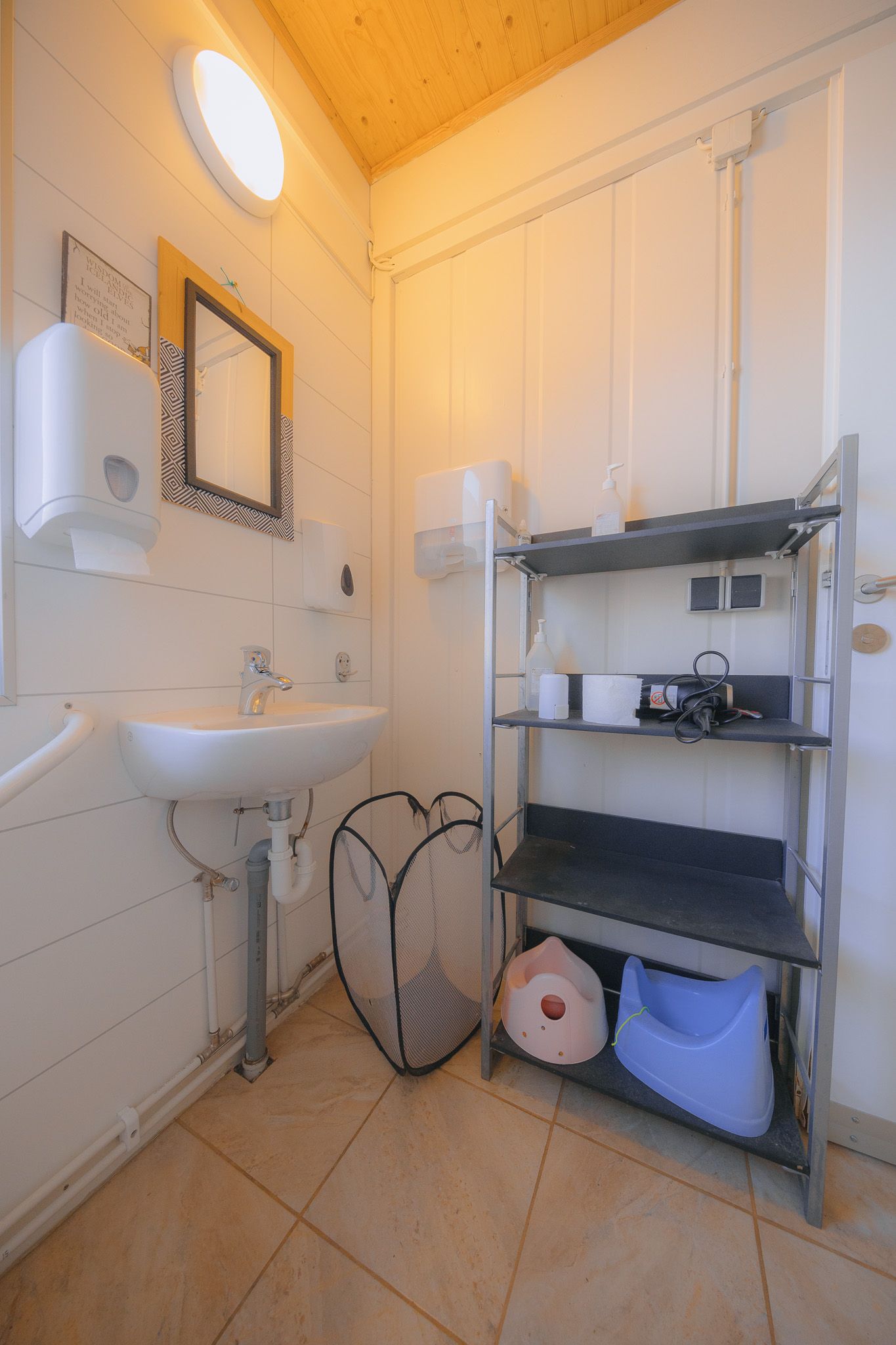 A small bathroom with a sink, a round mirror, and a metal shelving unit. Items include soap dispensers, a trash basket, and children’s toilets.