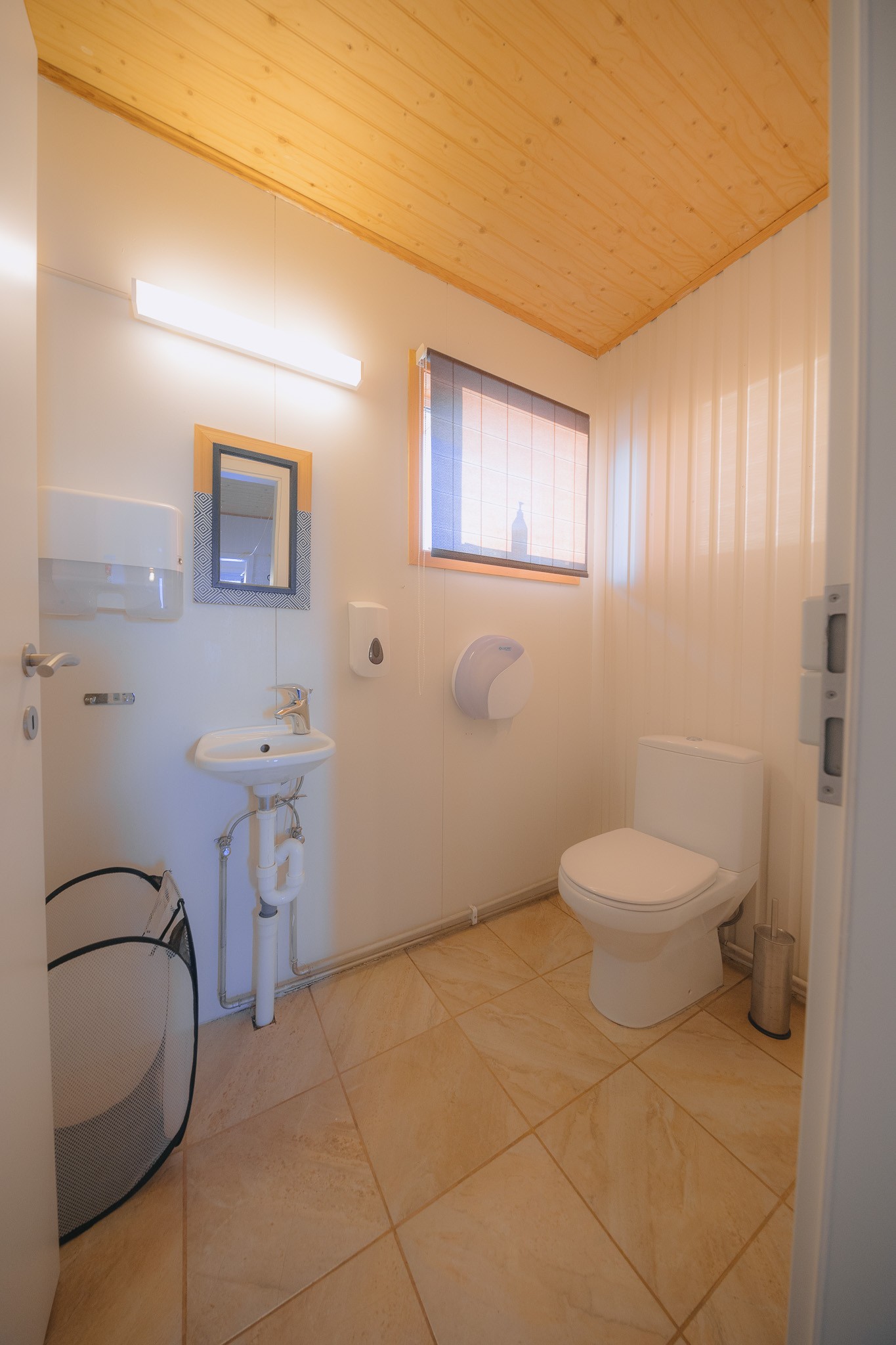 Compact bathroom with beige tile floor and wooden ceiling. Features a small sink with a mirror, a toilet, and a window letting in soft light.