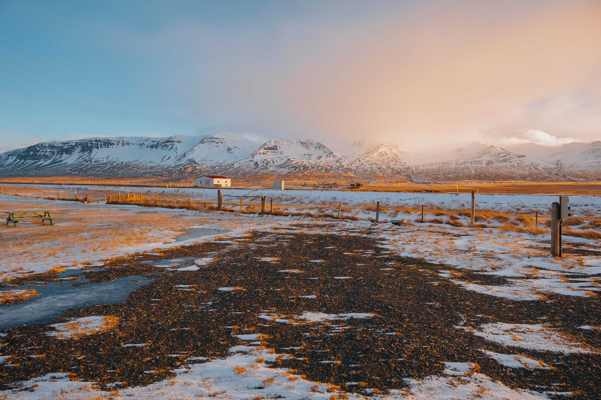 Snow-dusted field with scattered patches of earth in foreground, leading to distant snowy mountain range.