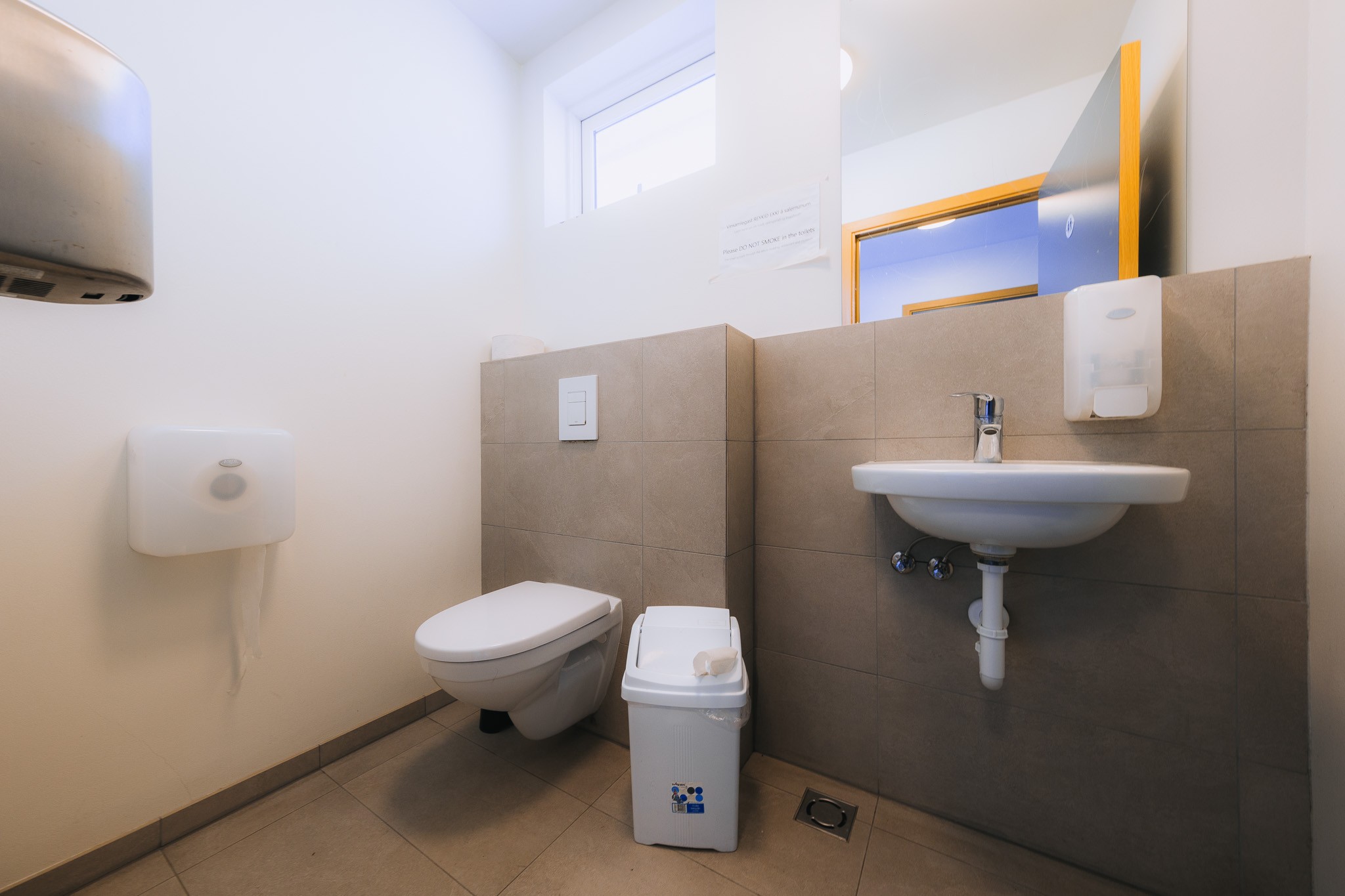Clean public restroom with tiled walls, featuring a wall-mounted toilet, sink with a soap dispenser, small bin nearby, and bright natural lighting.