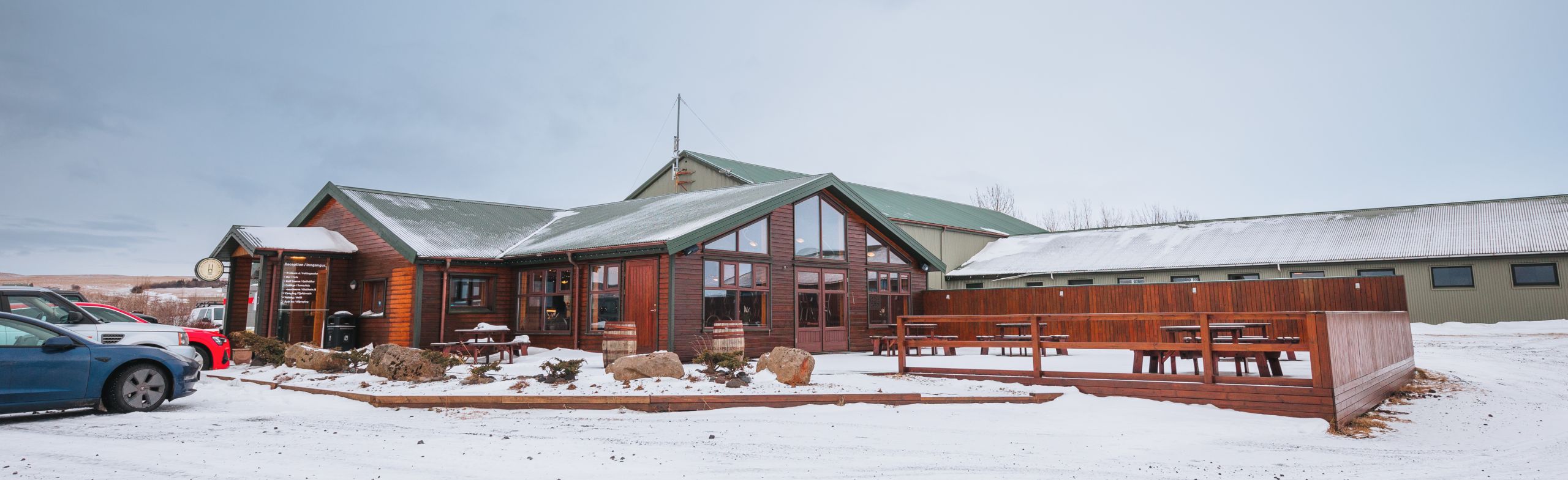 Snow-covered landscape with a rustic wooden building featuring green roofs. Picnic tables and cars are visible outside.