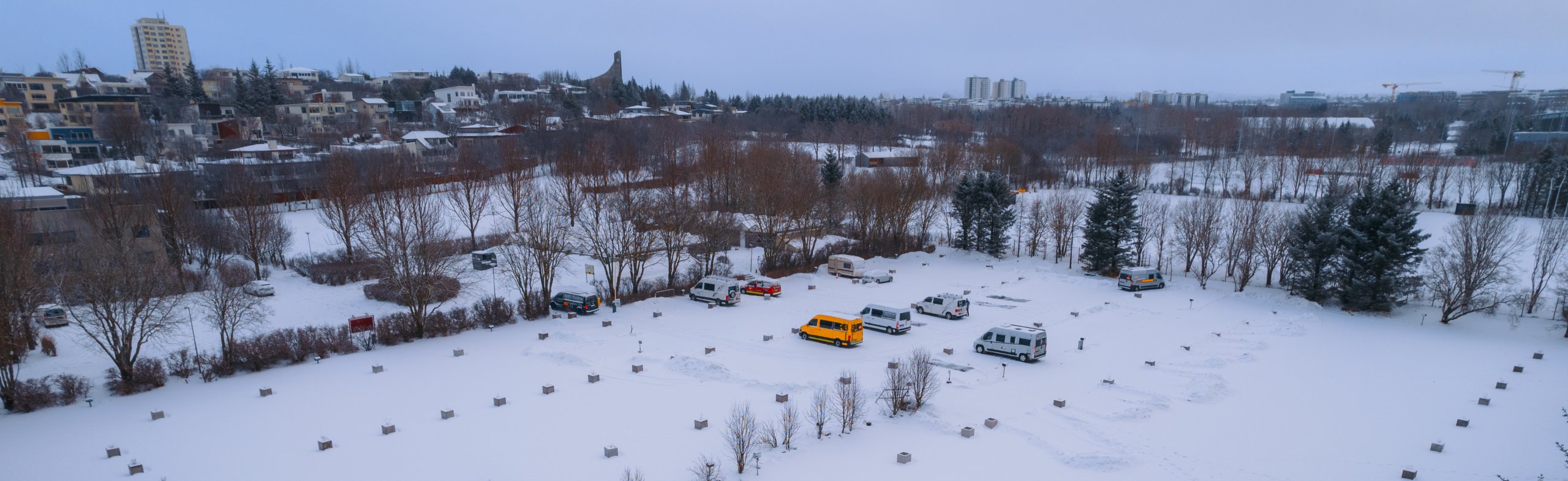 Snow-covered landscape with scattered vehicles, including a bright yellow van, and bare trees. Buildings and cloudy sky in the background.