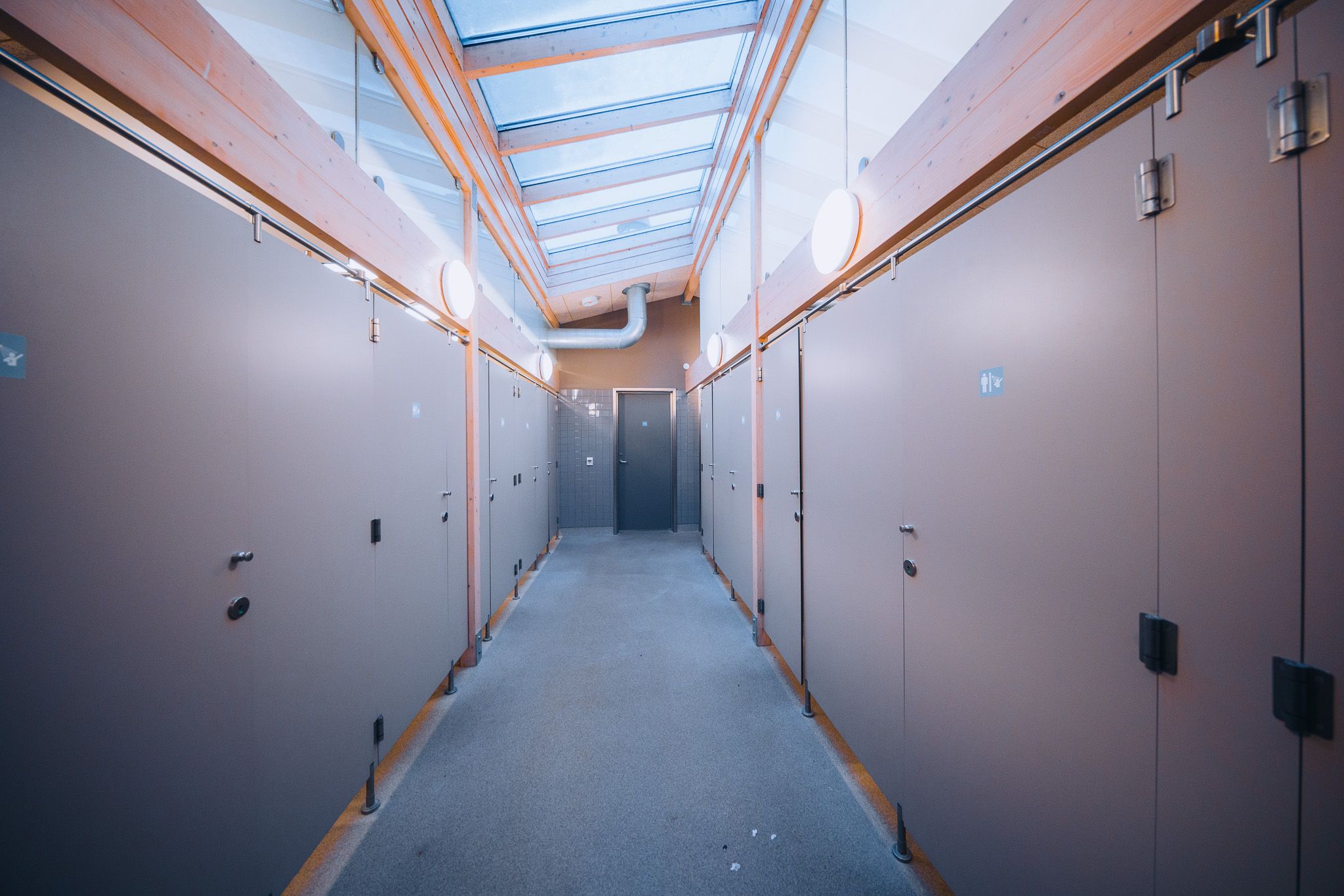 A narrow corridor with light gray walls and doors, illuminated by round ceiling lights and skylights.