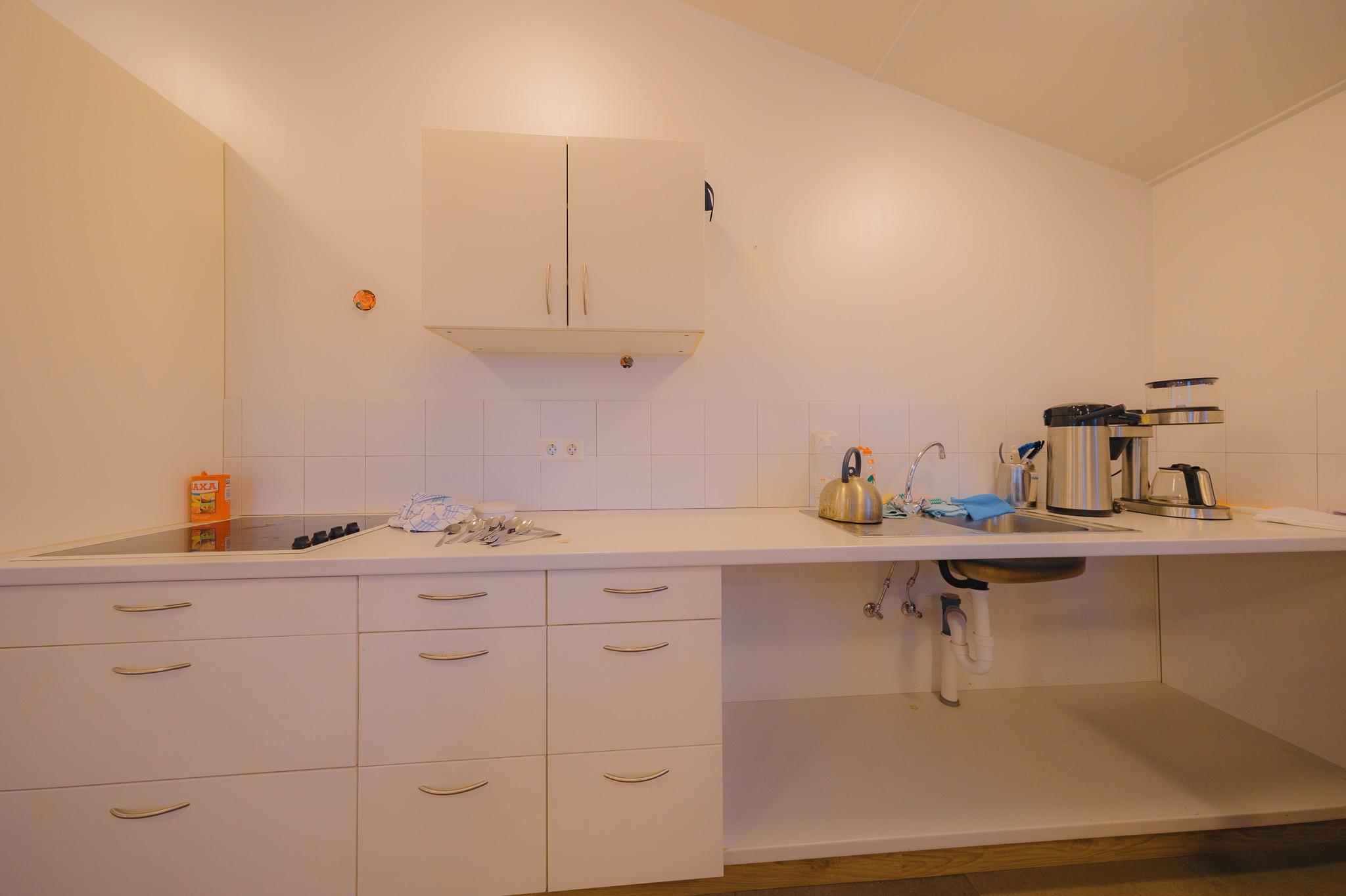 A clean, minimalist kitchen features white cabinets, a stovetop, a sink with a kettle, and a coffee maker.