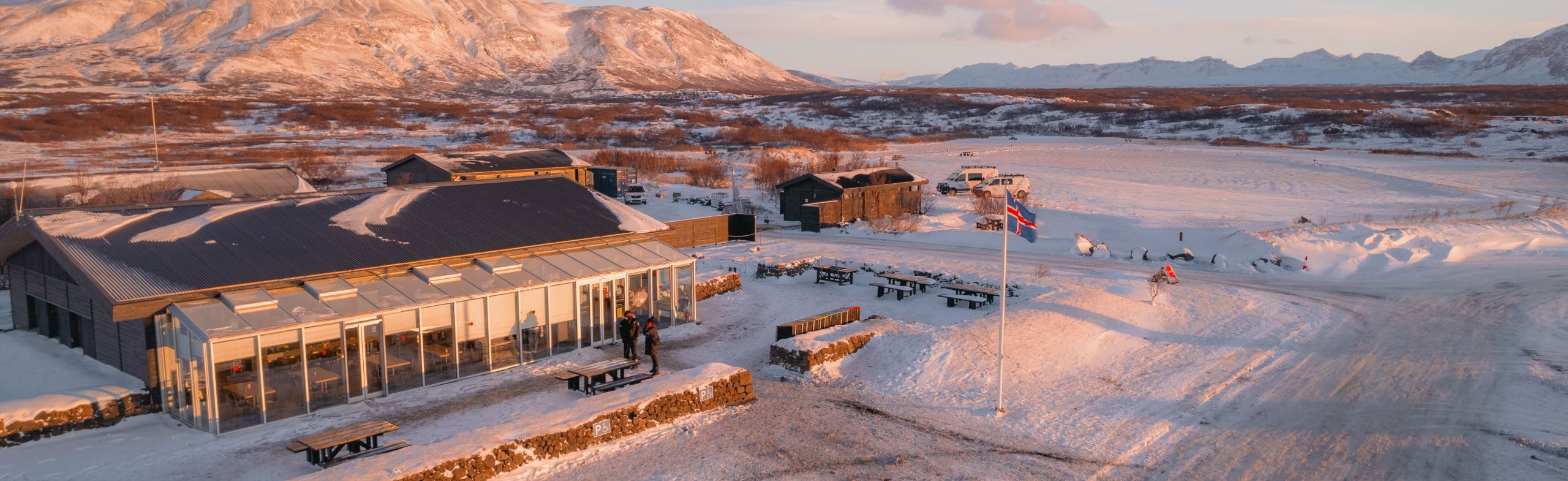 A snow-covered landscape at sunset features a building with large windows, a flagpole, and mountains in the background.